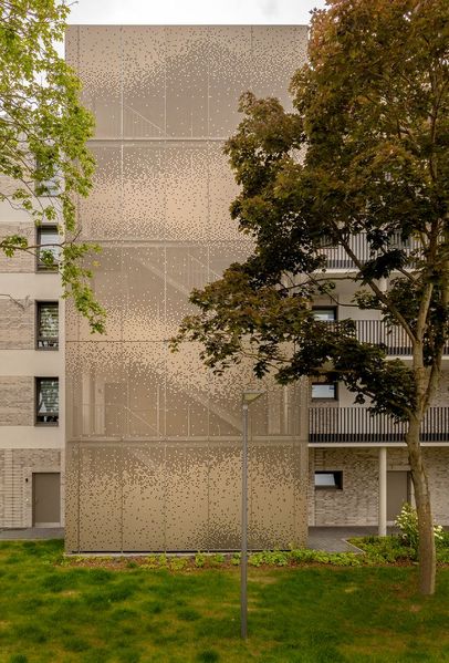 Außenansicht eines Wohngebäudes am Obentrautpark in Berlin. Ein markantes, mehrstöckiges Treppenhaus ist vollständig mit gold-bronzefarbenen Lochblech-Paneelen verkleidet. Die semitransparente Metallhülle lässt die dahinterliegenden Treppenläufe schemenhaft erkennen und bildet einen warmen, edlen Kontrast zu den umgebenden grünen Bäumen und der hellen Fassade des Hauptgebäudes.